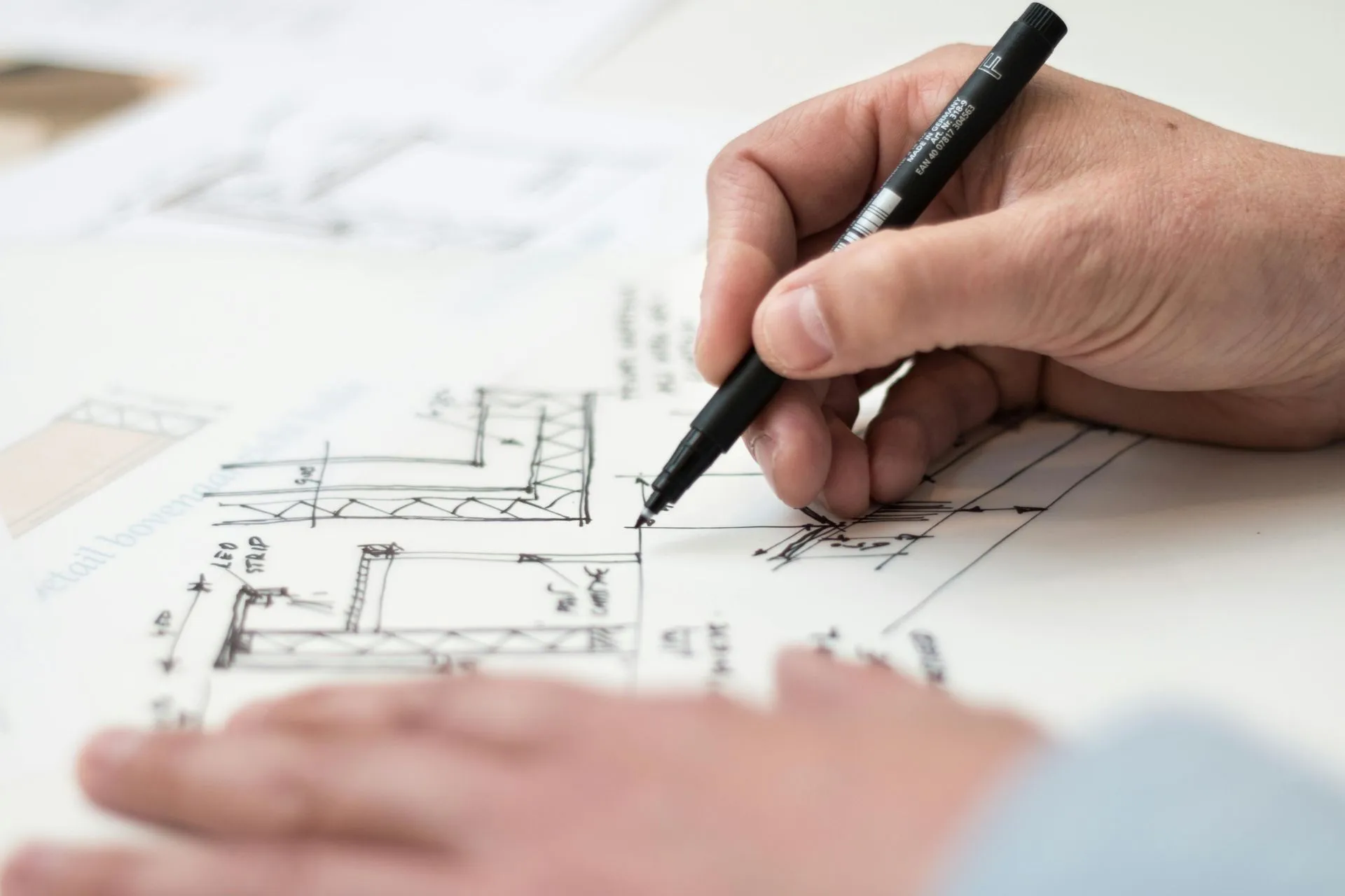 A hand holding a pen draws detailed architectural sketches on white paper, showing lines, measurements, and angles in a technical design.