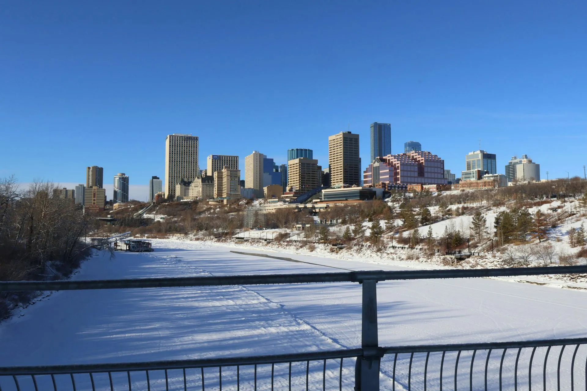 City skyline view with tall buildings under a clear blue sky, overlooking a snow-covered river and hilly landscape in the foreground.