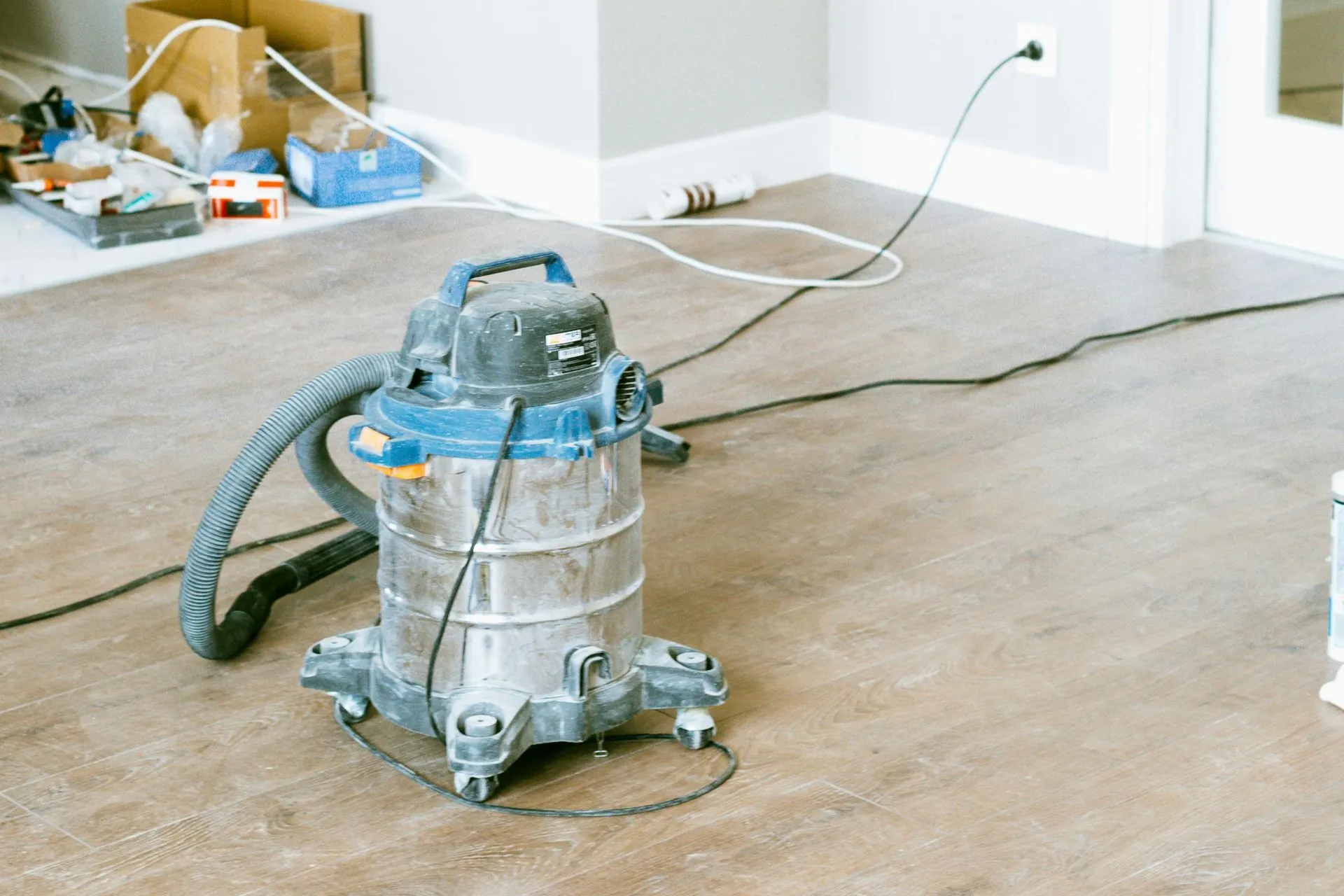 A dusty vacuum cleaner sits on a wooden floor in a home renovation setting. In the background, scattered tools and an electrical cord are visible.