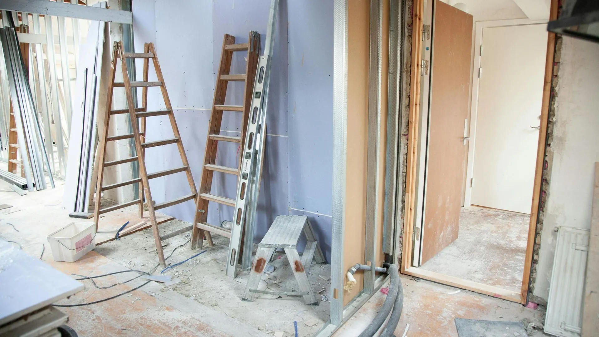 Room under renovation with wooden ladders, metal beams, pipes, and a partially open door. Dust and construction materials are scattered around.
