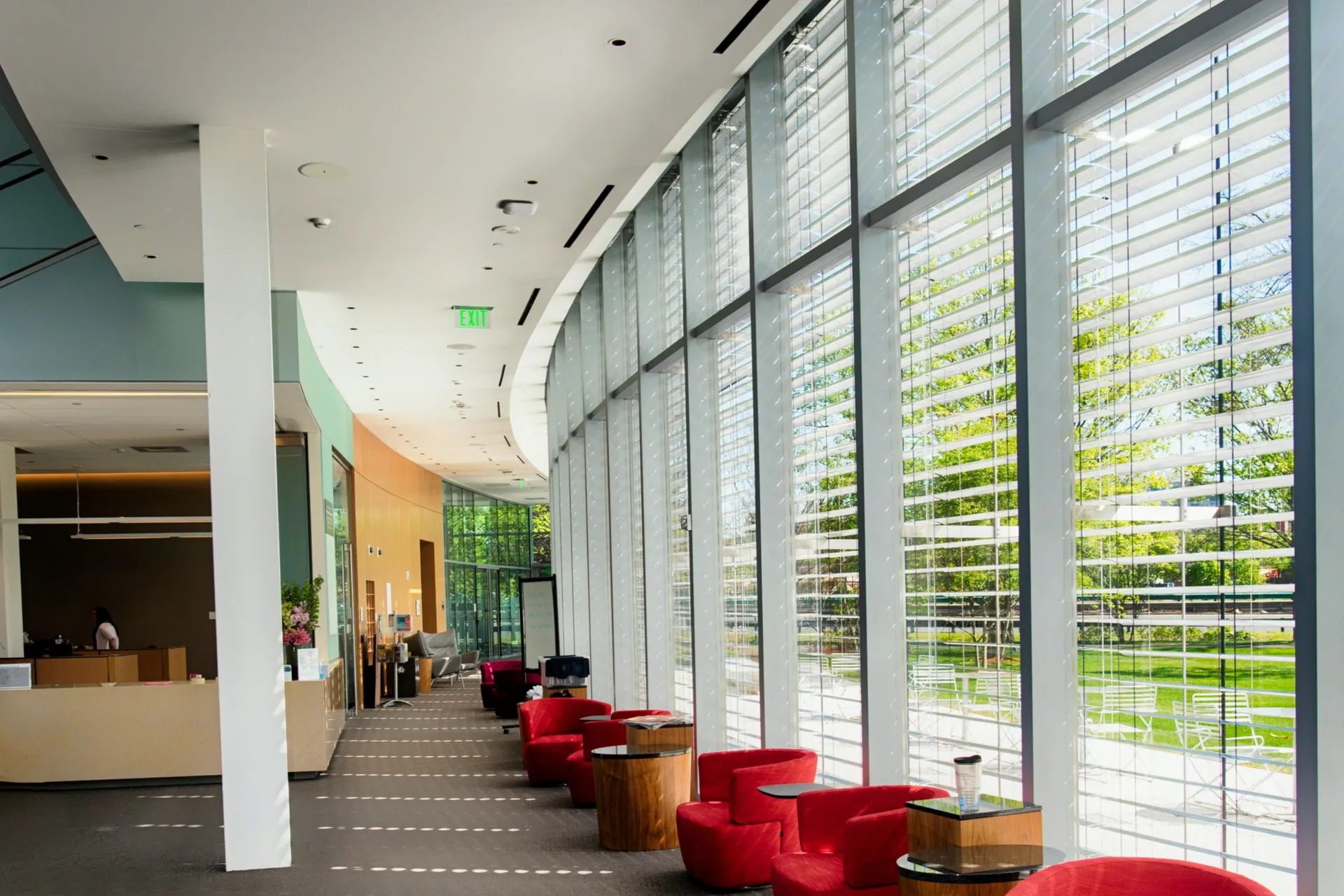 Modern lobby with large windows and horizontal blinds. Red chairs and small tables line one side. Sunlight filters through, casting patterns on