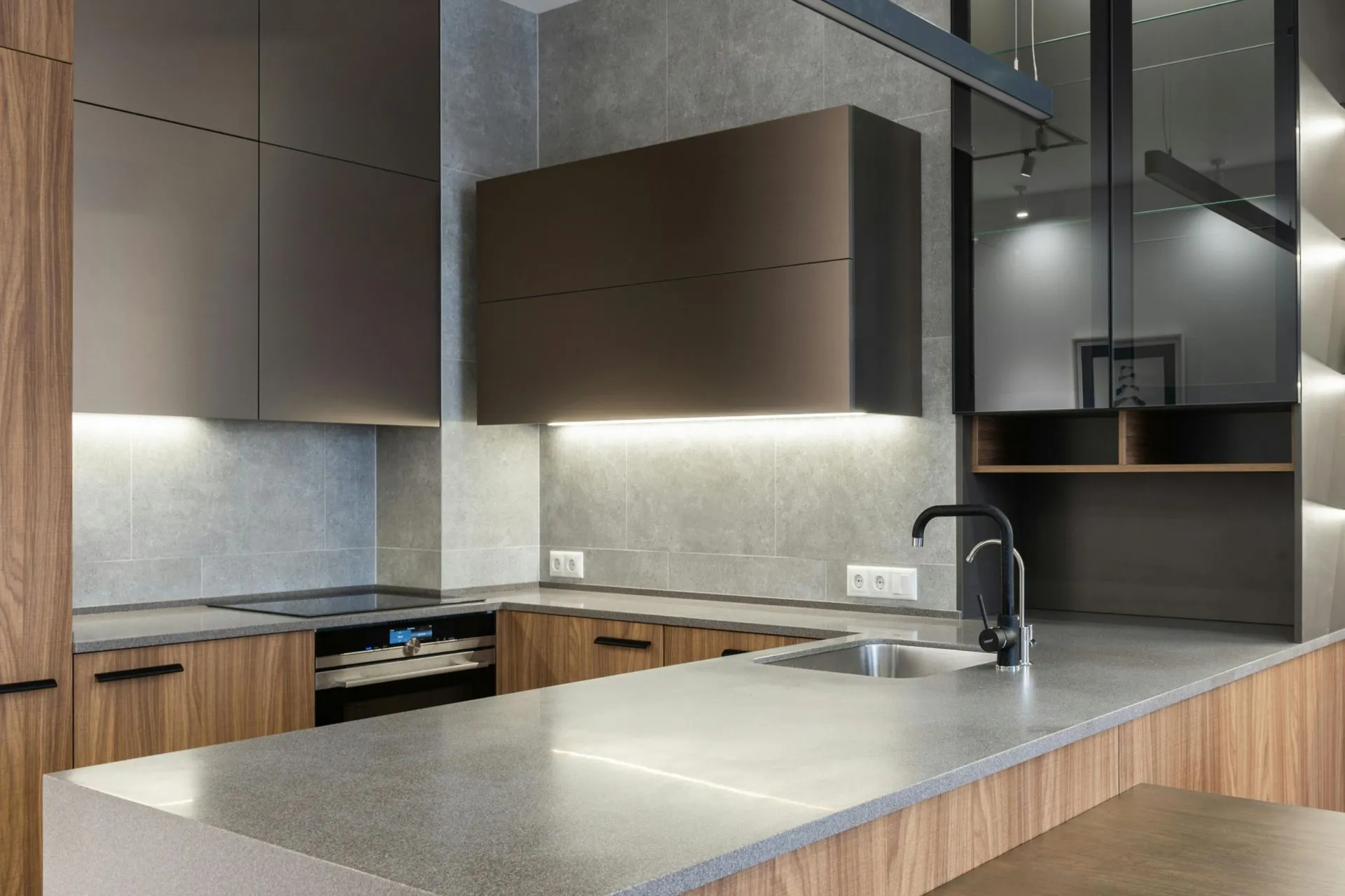 Modern kitchen with gray countertops, wooden cabinets, under-cabinet lighting, electric stovetop, stainless steel sink, and sleek black tap.