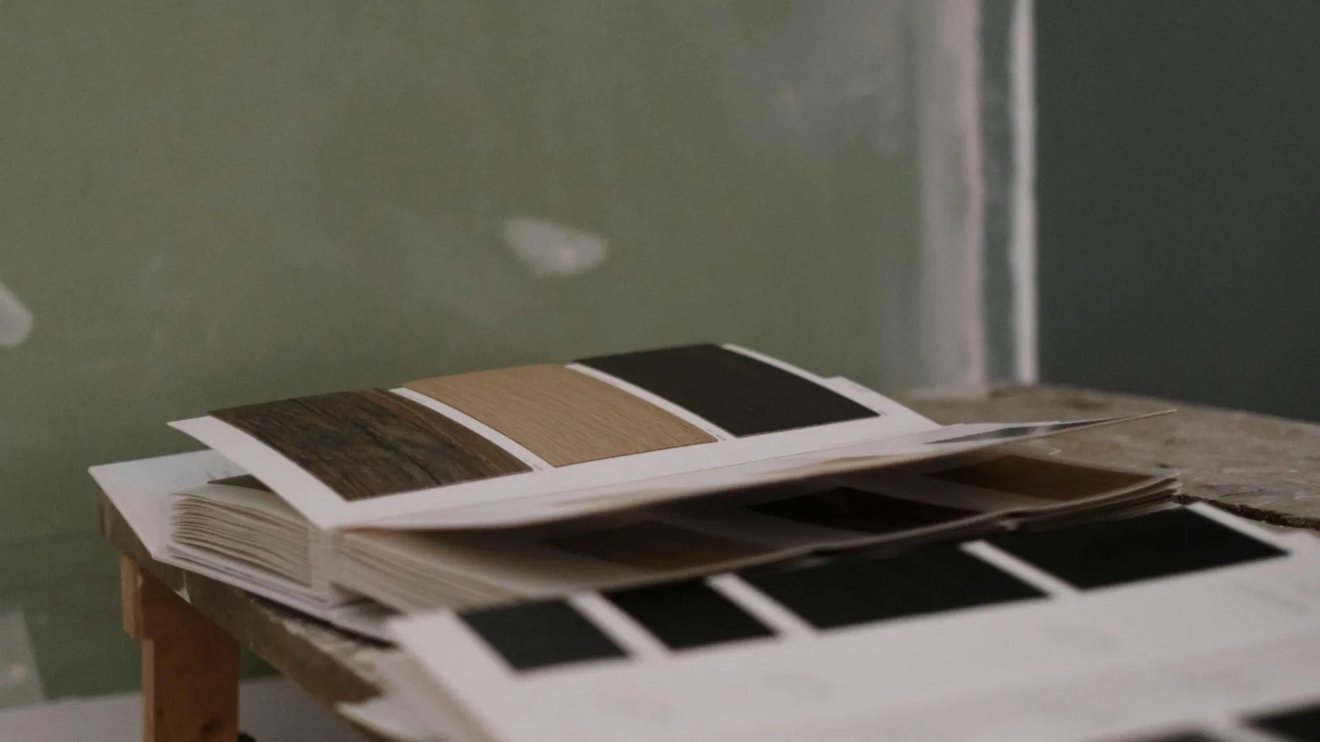 Open catalog on a wooden table showing fabric or material swatches in various shades, including brown and black, against a muted green background.