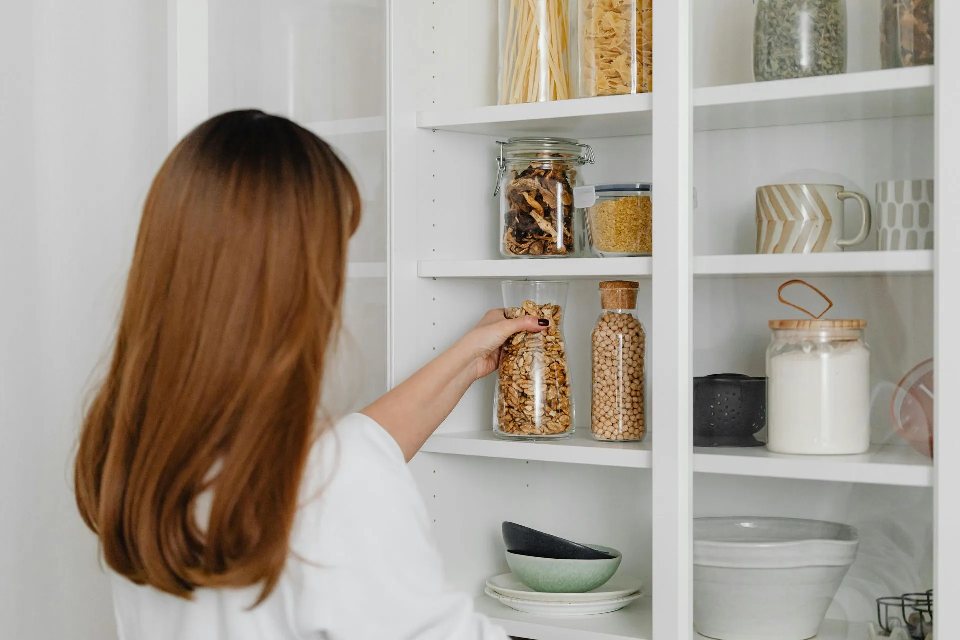 A woman with long brown hair organizes jars of food in a white pantry, including grains, pasta, and legumes, with bowls and mugs on the shelves.