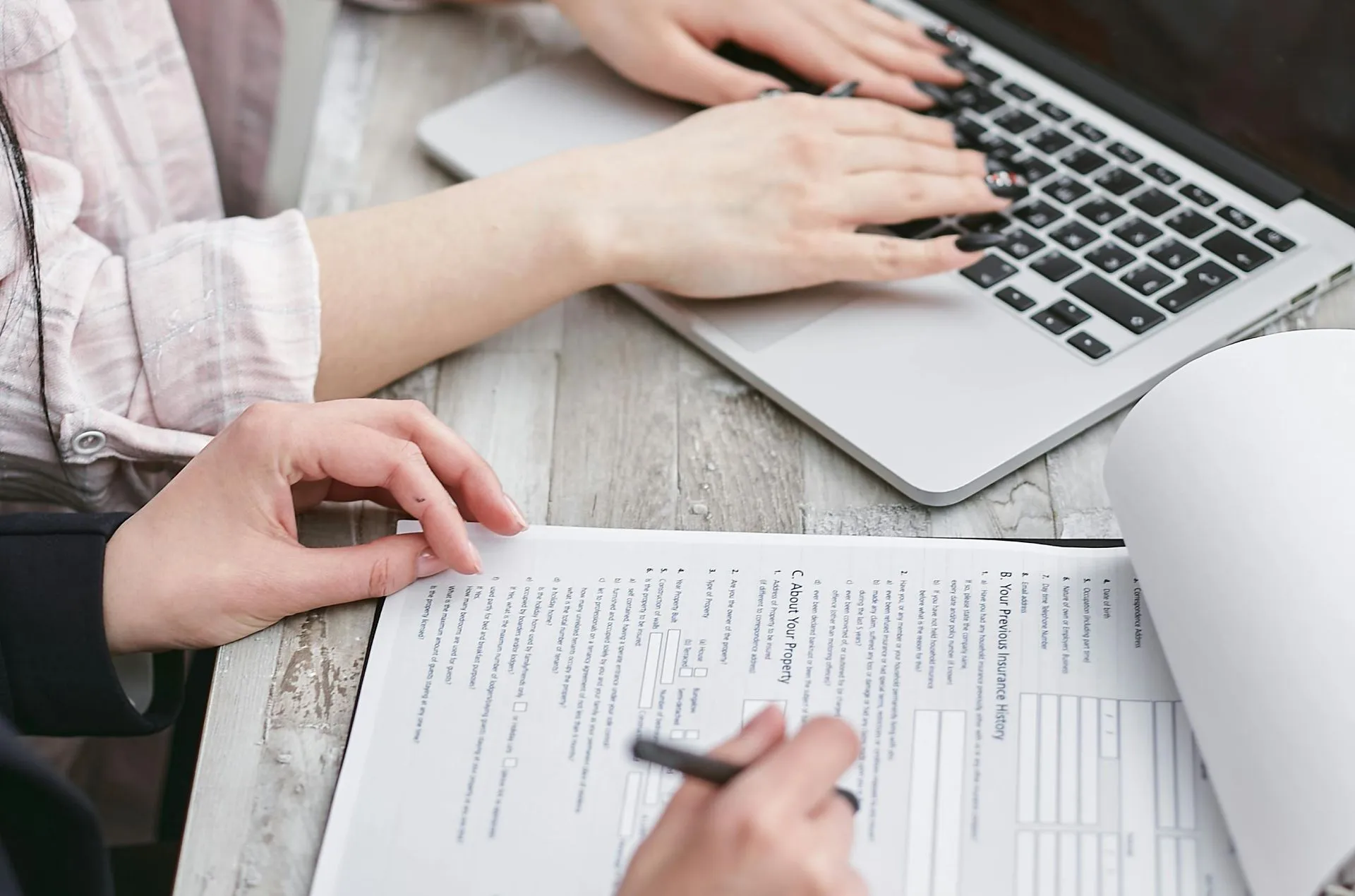 Hands typing on a laptop and filling out a paper form with a pen on a wooden table.