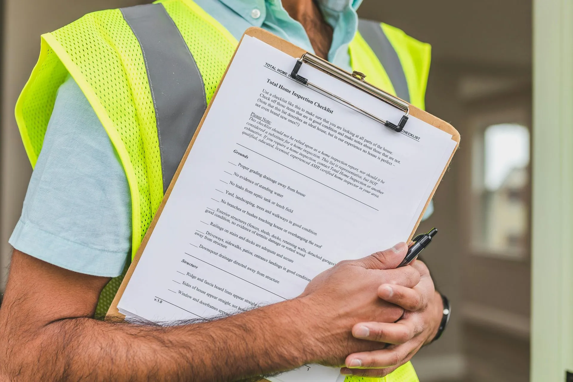 A person in a reflective vest holding a clipboard with a Total Home Inspection Checklist, with pen in hand, standing indoors.