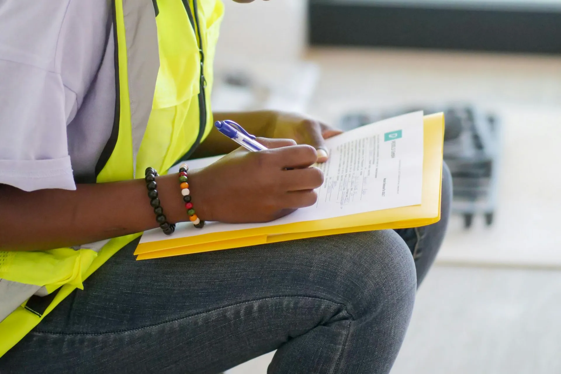 Person in a yellow safety vest writing on a clipboard with a blue pen, wearing a colorful beaded bracelet, sitting on a knee.