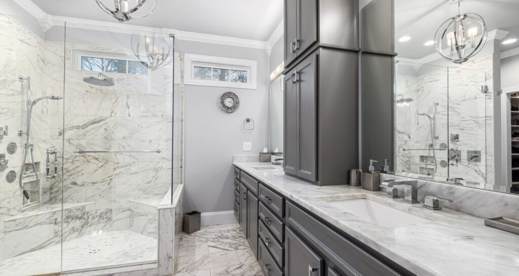 Modern bathroom with marble shower, large mirror, dual sinks, and gray cabinets. Chandeliers and a wall clock enhance the elegant design.